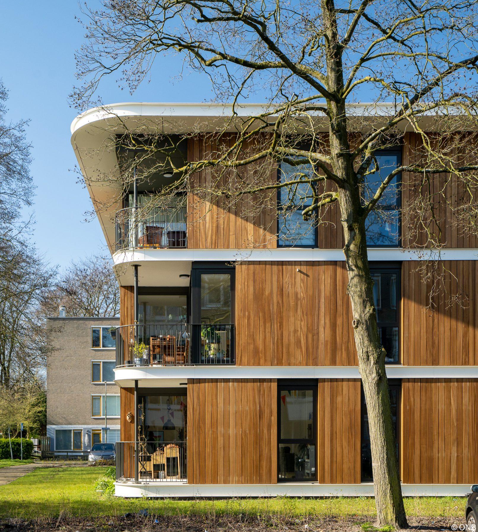 Tussen Utrechtse flatwoningen in een groene ruimte staat een modern houten woongebouw van drie bouwlagen, iets opgetild boven het landschap. Woonpaviljoen Camposdreef, CRKL architecten