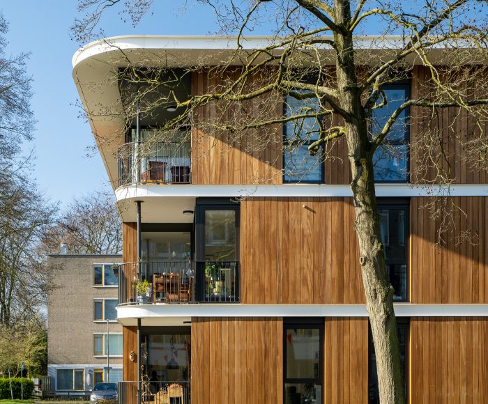 Tussen Utrechtse flatwoningen in een groene ruimte staat een modern houten woongebouw van drie bouwlagen, iets opgetild boven het landschap. Woonpaviljoen Camposdreef, CRKL architecten